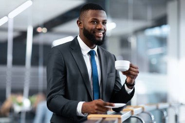 Bir işadamı modern bir ofiste dururken sağ elinde bir kahve fincanı ve solunda bir fincan fincan tutar. Etrafa bakarken gülümser, takım elbise giyer ve kravat takar..