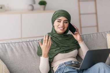 Başörtülü genç, olumlu Arap kadın evdeki kanepede oturuyor. Dizüstü bilgisayar kullanıyor ve akıllı telefonuyla konuşuyor. Neşeli görünüyor ve iş arkadaşlarıyla meşgul..