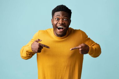 Çekici, genç, siyah bir adam sarı kazağını işaret ediyor. Mutlu, Afro-Amerikan milenyum adamı kıyafetlerinde bir şey gösteriyor. Mavi stüdyo arka planı, fotokopi odası.