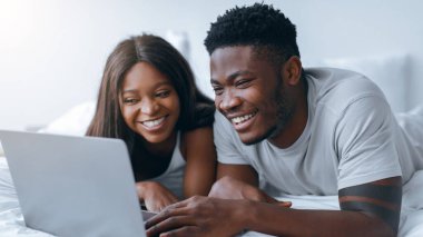 İki kişi bir yatakta oturup dizüstü bilgisayara odaklanmış. Gülümsüyorlar ve birbirlerine eşlik etmekten zevk alıyorlar. Güneş ışığı pencereden süzülür ve sıcak bir atmosfer yaratır..