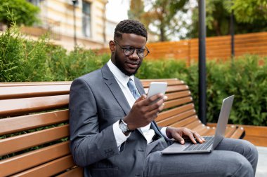 Genç Afrikalı Amerikalı iş adamı laptopuyla bankta oturuyor ve akıllı telefon kullanıyor, boş yer var. Modern dizüstü bilgisayar ve cep telefonu kullanan kendine güvenen bir zenci.