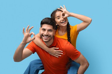 Bir kız ve bir oğlan parlak mavi bir arka plana karşı neşeli pozlar veriyorlar. Kız oğlanların sırtına oturuyor, hem gülüyor hem de el hareketleriyle eğleniyor..