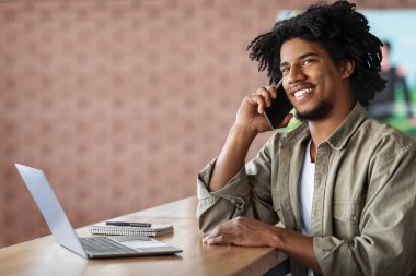 Gülümseyen, cep telefonuyla konuşan ve kafede dizüstü bilgisayarla çalışan Afro-Amerikan genç adam, keyifli mobil konuşmalar yapıyor ve bilgisayar kullanıyor, uzaktan kumandayla eğleniyor, boşluğu kopyalıyor.