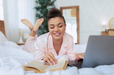 Milenyum siyahi bayanı dizüstü bilgisayarla yatakta uzanıyor, kitap okuyor ve bilgisayarla online ders alıyor, evden uzaktan çalışıyor ve gülümsüyor.