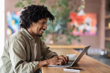 Neşeli, Afro-Amerikan kıvırcık erkek kahve fincanı ve telefonuyla dizüstü bilgisayarda yazı yazıyor. Uzaktan çalışmada bağımsız yüklenici, iş yerinde dijital göçebe, blog yazarı ve sohbet