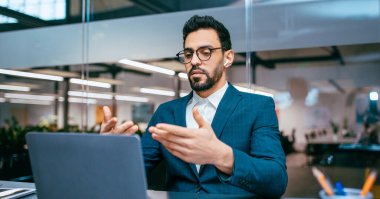 Modern bir ofiste bir adam masada oturuyor. Resmi kıyafetler giyiyor ve konuşurken ve el hareketi yaparken bir dizüstü bilgisayar kullanıyor. Ofiste bir sürü fabrika var..
