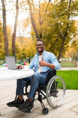 Engelliler için internet işi. Tekerlekli sandalyedeki gülümseyen özürlü adam dizüstü bilgisayarla çalışıyor, sonbahar parkındaki kafede baş parmak hareketi gösteriyor, uzaktan iş öneriyor, kopyalama alanı öneriyor.