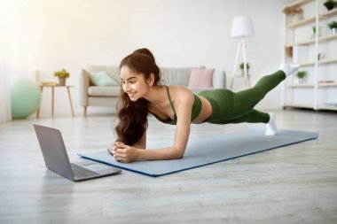 Güçlü Hintli kadın dirsek tahtası üzerinde duruyor, bacağını spor minderinin üzerine kaldırıyor, evde dizüstü bilgisayarında video dersi takip ediyor. Doğulu genç bayan vücut kaslarını çalıştırıyor, ev içi spor yapıyor.