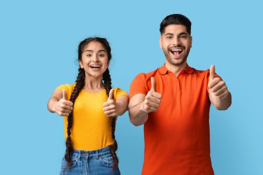 İki genç yetişkin yan yana, gülümseyerek başparmak kaldırıyor. Heyecanlı ve neşeli görünüyorlar, parlak mavi bir arka planda bir mutluluk anı sergiliyorlar..