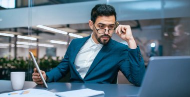 Takım elbiseli bir adam elinde tablet tutarken bilgisayarına odaklanmış bir şekilde ofis masasında oturuyor. Verileri ya da belgeleri analiz ediyor gibi görünüyor. Masanın üzerinde bir fincan ve bir not defteri var..