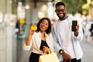 İnternetten alışveriş. Kredi kartı tutan heyecanlı Afro-Amerikan çiftin portresi ve kameraya siyah ekranlı cep telefonlarını gösteren alışveriş torbaları. Model için boş alan, dijital ödeme