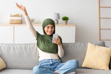 Heyecanlı genç Arap kadın rahat koltuğunda oturup akıllı telefonuyla mobil oyun oynuyor. Memleketindeki yeni oyun uygulamasının keyfini çıkarırken sevinçle bir zafer jesti yapıyor..