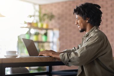 Modern kafenin iç kısımlarında dizüstü bilgisayarda yazı yazan mutlu, kıvırcık, genç bir erkek. Güneş ışığı, profil. Dijital göçebe, serbest çalışan, kapalı alanda çalışan, yeni normal ve teknoloji. Çalış ve uzaktan çalış