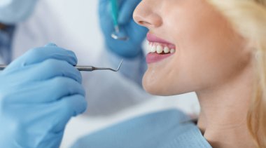 Gülümseyen genç sarışın kadın ve dişçi elleri mavi hırsız eldivenleri, diş aletleri, yan görünüş, panorama. Tanımlanamayan kadın hasta diş hekimliğini ziyaret ediyor, düzenli kontrol yaptırıyor.