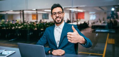 Takım elbiseli bir adam dizüstü bilgisayarı olan bir masada oturuyor ve başparmağıyla gülümsüyor. Ofiste gün içinde bitkiler ve parlak bir çalışma alanı var..