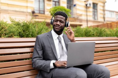 Kablosuz kulaklık takan, laptop kullanan ve kapalı gözlerle müzik dinleyen Afro-Amerikan iş adamı büro merkezinin yanındaki bankta oturuyor. Mutlu siyahi girişimciler, müzik listesinin keyfini çıkarın.