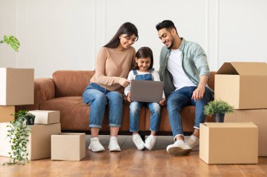 Yeni eve taşındıktan sonra dizüstü bilgisayarı koltukta otururken mutlu aile ilişkileri. Gayrimenkul, Ev Sahipliği ve Taşınma Konsepti. İnsanlar karton kutuların arasında oturup, ekranı işaret ederek internete bakıyorlar.
