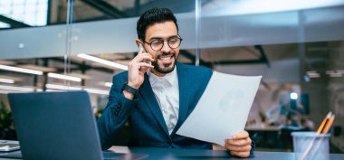 Takım elbiseli bir adam masada oturuyor, telefonla konuşuyor ve bir kağıt parçasına bakıyor. Masada bir dizüstü bilgisayar ve ofis malzemeleri görülebilir. Doğal ışık pencereden içeri giriyor..