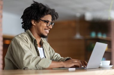 Gözlüklü, Afro-Amerikan kıvırcık saçlı genç adamın laptopta, kafenin içinde, profili olan bir masada çalışmasına sevindim. Bağımsız müteahhit çalışma alanında çalışıyor, iş yerinde modern teknolojiyle çalışıyor.
