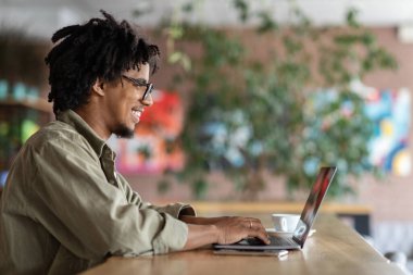 Blog Kavramı. Sosyal ağlara içerik yüklemek için dizüstü bilgisayar kullanan neşeli genç siyahi adam, Milenyum Afrikalı Amerikalı blogcu kahve dükkanında otururken bilgisayarda yazı yazıyor, Boşluğu Kopyala