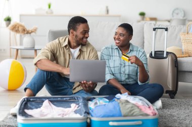 Keyifli Siyah Gezginler çift dizüstü bilgisayar ve kredi kartı kullanıyor tatil rezervasyonu yapıyor ve evde bavullarıyla oturan seyahat biletleri alıyor. Turistlerin Uçuş Defteri Bilgisayarla. Seçici Odaklanma