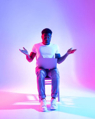 Şok olmuş Afro-Amerikalı bir adam bir sandalyeye oturup, laptopuna ağzı açık bir şekilde bakıyor. Canlı neon ışıklarıyla aydınlanıyor ve fantastik bir online anlaşmayla heyecanını gösteriyor..