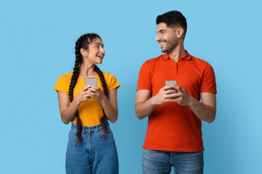 İki arkadaş yan yana durur, telefonlarını tutarken birbirlerine gülümserler. Parlak renklerle giyinmişler, canlı bir ortamda mutluluk yayıyorlar..