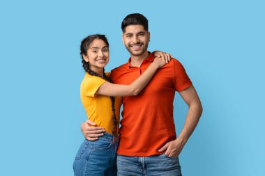 İki genç yetişkin birbirine yakın duruyor, gülümsüyor ve sarılıyorlar. Erkek turuncu gömlek ve kot pantolon giyerken, kadın sarı gömlek ve kot şort giyiyor..