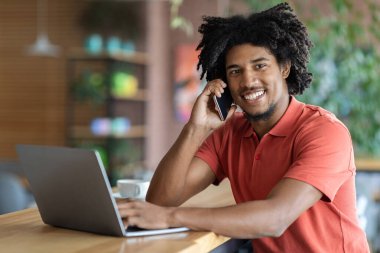 Modern Girişimci. Kafede masada otururken cep telefonuyla konuşan ve dizüstü bilgisayar kullanan mutlu siyahi iş adamı, kafeteryada bilgisayarla uzaktan çalışan neşeli Afrikalı Amerikalı adam,