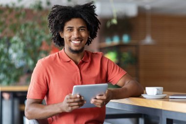 Neşeli milenyum siyah kıvırcık adam blogcusu modern kafenin içinde bir fincan kahveyle tablet üzerinde yazı yazıyor. Sosyal ağlar, blog ve sohbet, uzaktan çalışma ve çalışma. İş, uygulama ve aygıt