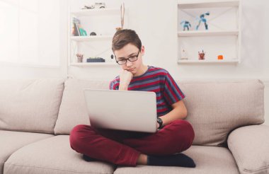 Bored boy with computer on sofa at home. Child engrossed in internet, typing on keyboard