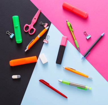 Çeşitli kırtasiye malzemeleri titizlikle siyah, pembe ve mavi bir yüzeye yerleştirilmiştir. Kalemler, kalemler, makas ve ataçlar, canlı bir çalışma alanı gösteren eşyalar.