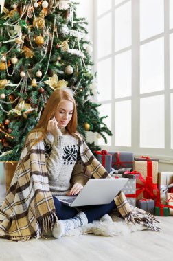 Cep telefonuyla konuşan genç bir kadın ve rahat bir Noel 'de dizüstü bilgisayarla alışveriş yapan bir kadın. Kız süslenmiş çam ağacının altında hediyelerin arasında oturuyor. Xmas 'a hazırlanmak, kış satışlarında hile yapmak, kopyalama alanı