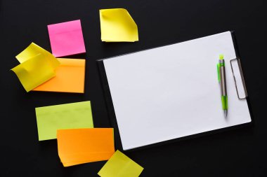 Pembe, turuncu ve yeşil renkli notlar siyah bir yüzeyde boş bir not defteri ve bir kalem etrafına saçılmış, beyin fırtınası veya planlama faaliyetleri için canlı bir çalışma alanı yaratıyor..