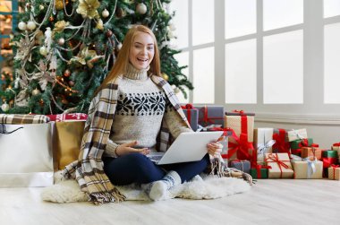 Dizüstü bilgisayarı olan mutlu kız süslü Noel ağacı arka planında oturuyor. Kadın internetten tebrik e-postası almış. Kış indiriminde büyük indirim varmış. Tatil atmosferi, boşluğu kopyala
