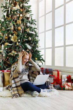 Dizüstü bilgisayarı olan mutlu kız süslü Noel ağacı arka planında oturuyor. Kadın internetten tebrik e-postası almış. Kış indiriminde büyük indirim varmış. Tatil atmosferi, boşluğu kopyala