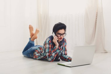 Laptop ile halı üzerinde yalan, mobil, cep telefonuyla konuşurken bilgisayar gözlüklü genç kız. Hipster öğrenci, serbest meslek sahibi kadın evde. Bilgisayar ile rahatlayın. Dizüstü bilgisayar ile kız. Yumuşak sesi, yüksek anahtar.