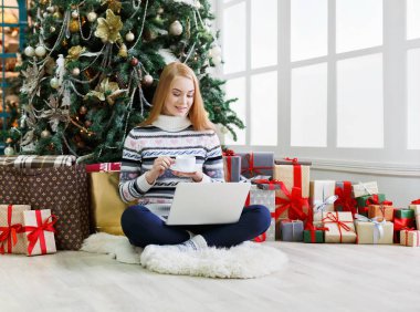 Yılbaşında internetten kahve alışverişi yapan genç bir kadın. Bir sürü hediye arasında süslenmiş köknar ağacının altında oturan kız. Xmas 'a hazırlanmak, kış satışlarında hile yapmak, kopyalama alanı