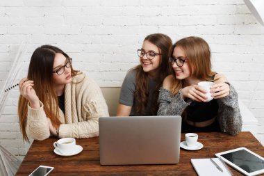 Üç arkadaş bir kafede bir masada oturuyorlar. Laptop kullanıyorlar ve kahve içiyorlar. Bir arkadaş notlar yazarken, diğerleri gülüyor ve konuşuyor..