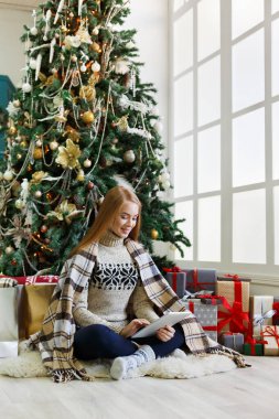 Genç bir kadın, rahat bir Noel 'de online tablet alışverişi yapıyor. Paketli hediyelerin arasında süslü çam ağacının altında oturan kız. Xmas 'a hazırlanmak, kış satışlarında hile yapmak, kopyalama alanı