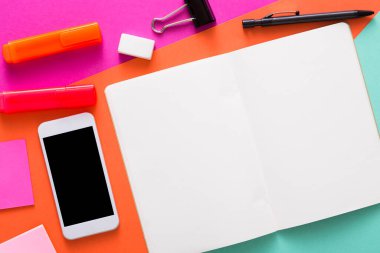 Canlı bir çalışma alanı açık bir boş defter, bir akıllı telefon, fosforlu kalemler, yapışkan notlar ve renkli bir arkaplana saçılmış bir silgi içerir. Kurulum beyin fırtınası ve yaratıcılık için ideal..