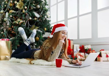 Noel Baba şapkalı mutlu kız, arkadaşlarıyla Noel 'de bilgisayarda görüntülü sohbet ediyor. Evdeki hediye kutuları ve paketlerin arasında süslü çam ağacının altında yatan genç kadın..