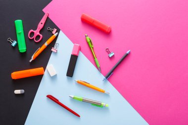 Renkli bir zemin üzerinde canlı bir kırtasiye malzemesi yatar. Kalemler, keçeli kalemler, makas ve klipler pembe, mavi ve siyah yüzeylere yayılmış, yaratıcı kullanıma hazır..