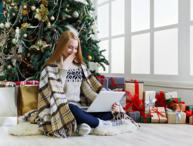 Dizüstü bilgisayarı olan mutlu kız süslü Noel ağacı arka planında oturuyor. Kadın internetten tebrik e-postası almış. Kış indiriminde büyük indirim varmış. Tatil atmosferi, boşluğu kopyala