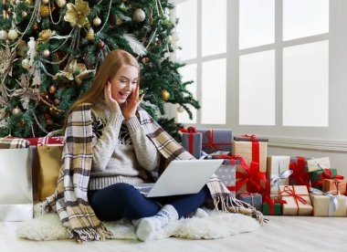 Dizüstü bilgisayarı olan mutlu kız süslü Noel ağacı arka planında oturuyor. Kadın internetten tebrik e-postası almış. Kış indiriminde büyük indirim varmış. Tatil atmosferi, boşluğu kopyala