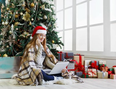 Cep telefonuyla konuşan genç bir kadın ve rahat bir Noel 'de dizüstü bilgisayarla alışveriş yapan bir kadın. Kız süslenmiş çam ağacının altında hediyelerin arasında oturuyor. Xmas 'a hazırlanmak, kış satışlarında hile yapmak, kopyalama alanı