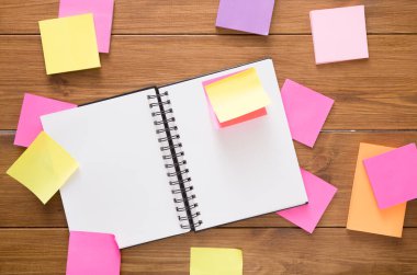 Boş defter, çeşitli renklerde parlak yapışkan notlarla çevrili ahşap bir masanın üzerinde yatıyor. Bu ayar, kişisel çalışma alanındaki beyin fırtınası veya planlama faaliyetlerini önerir.