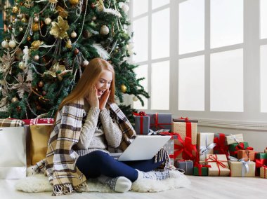 Dizüstü bilgisayarı olan mutlu kız süslü Noel ağacı arka planında oturuyor. Kadın internetten tebrik e-postası almış. Kış indiriminde büyük indirim varmış. Tatil atmosferi, boşluğu kopyala