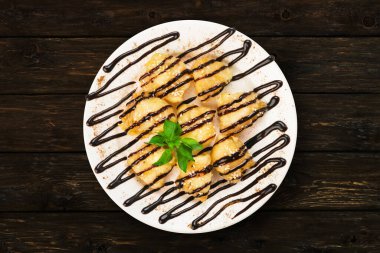 Koyu ahşap bir masada bir tabak tatlı duruyor. Tatlı çikolata sosuyla kaplanmış ve taze nane yaprağıyla süslenmiş. Tabakta birkaç ısırık büyüklüğünde porsiyon var..