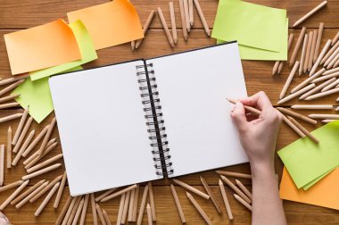 Bir insan eli, renkli kalemler ve ahşap bir masanın üzerindeki yapışkan notlarla çevrili açık bir çizim defterinin üzerinde bir kalem tutar. Ayarlar yaratıcı beyin fırtınası için düzenlendi..